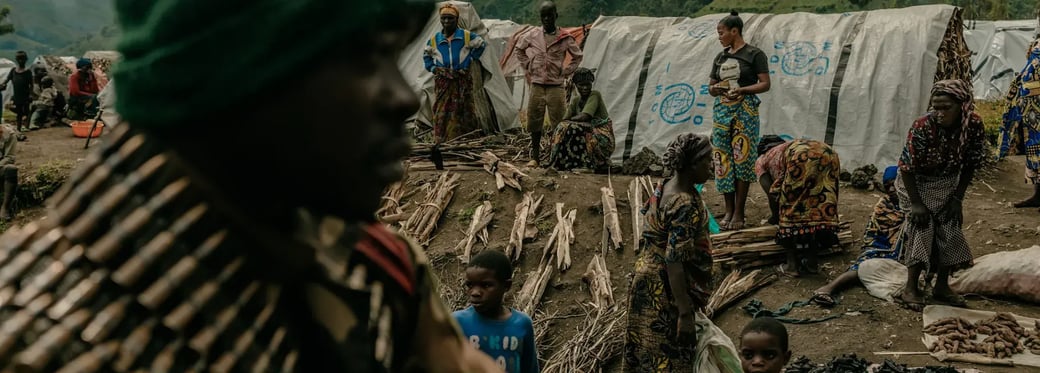 A soldier in a green beanie and bandolier stands in the foreground, with displaced people in makeshift shelters in a mountainous rural area.