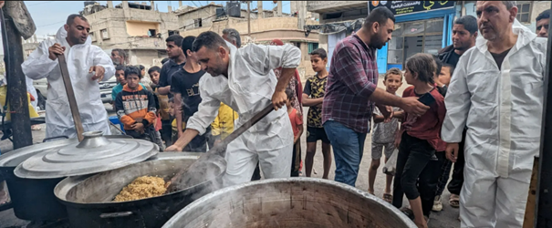 Workers in white overalls stir large vats of food in a community kitchen in Deir al-Balah, Gaza, with people and buildings in the background.