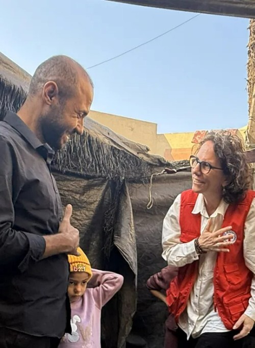 Yasmine Praz Dessimoz, ICRC Director of Operations,wearing a red vest and glasses shares a warm, smiling conversation with a man in a black shirt. They are standing in an outdoor setting next to a makeshift shelter. A young child wearing a yellow beanie is visible between them.