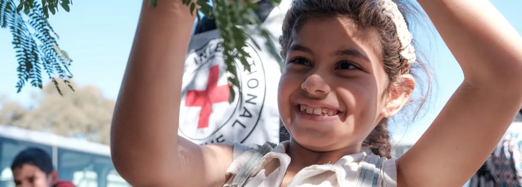 Batoul plays with a tree branch, smiling, with blue sky and a red cross in the background.