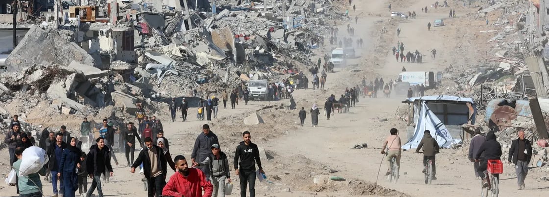 A wide, dusty road filled with people walking, cycling, and pulling carts through a landscape of immense destruction. To the left, buildings have been reduced to massive piles of concrete rubble and twisted rebar. The road stretches far into the distance, where more people and a few white vehicles are visible through the haze of dust.