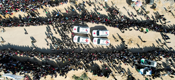 ICRC ambulances drive through dense crowds in a conflict zone, flanked by thousands lining both sides of a dusty road.