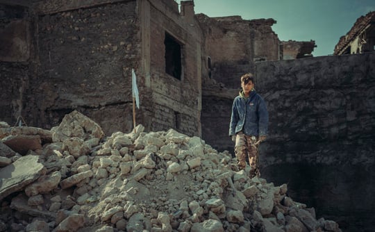 Photo of child in urban warfare setting standing on a pile of rubble