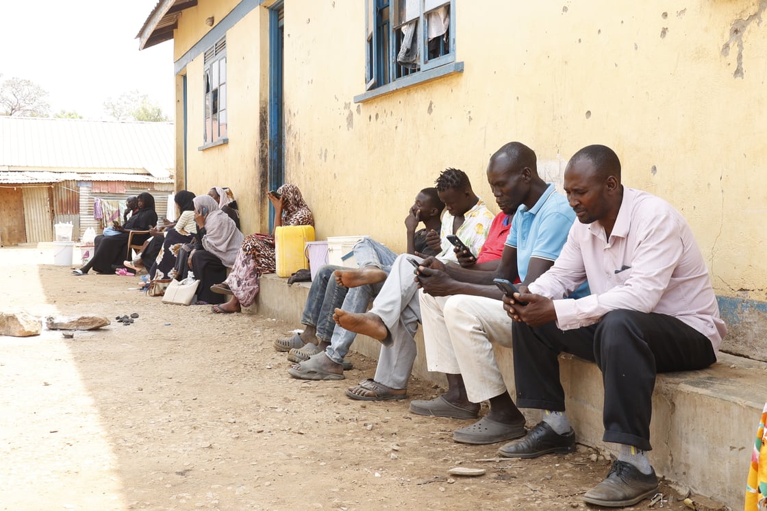 Several men and women sit in a row outside a building in a displacement camp, using their mobile phones to access digital family-tracing services provided by the ICRC.