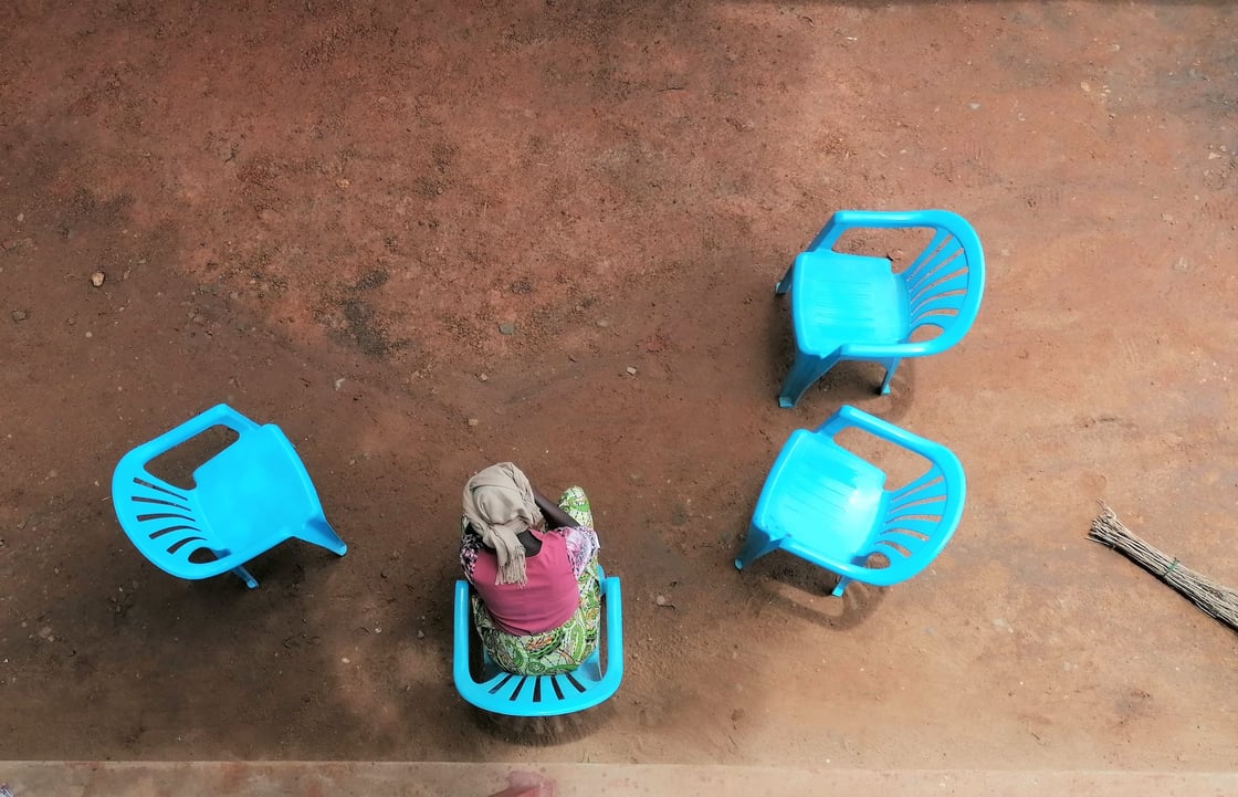 Aerial view of a woman seated alone on a blue plastic chair, surrounded by three empty ones. Her face remains hidden from view.