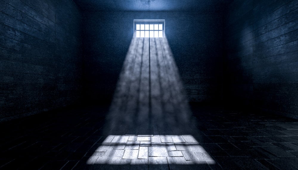 A dark prison cell with a single barred window casting a narrow beam of light onto the cold, empty floor.