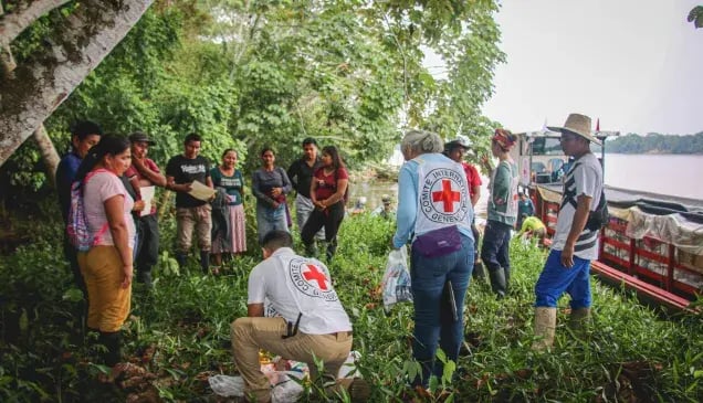 ICRC workers distribute aid packages to people gathered under trees near the Caquetá River, with a boat loaded with supplies docked nearby.